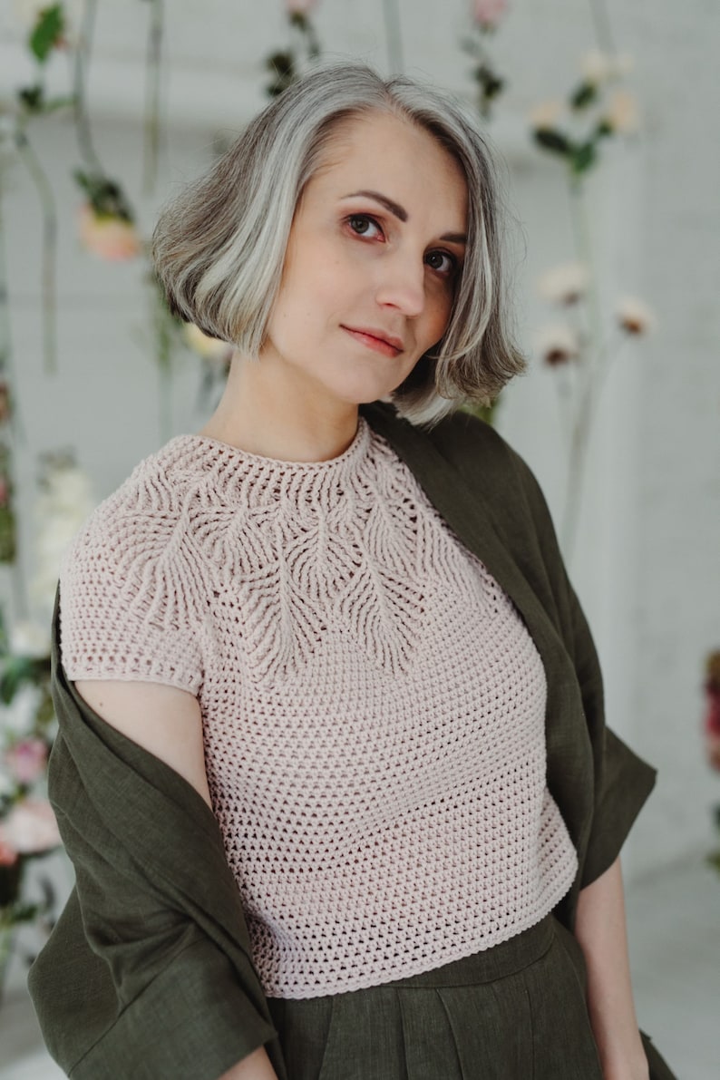 May include: A woman with short, gray hair is wearing a light pink crocheted top with a delicate leaf pattern. She is also wearing a dark green linen jacket and matching pants.