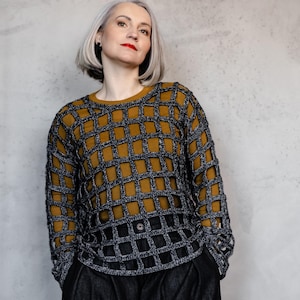 May include: A mustard yellow and gray open-weave sweater with a grid pattern. The sweater is worn over a mustard yellow top and paired with black pants. The model has short gray hair and red lipstick.