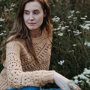May include: A woman wearing a light brown crocheted sweater and blue jeans sits in a field of white wildflowers.