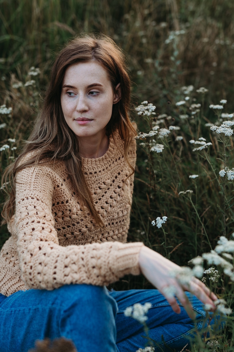May include: A woman wearing a light brown crocheted sweater and blue jeans sits in a field of white wildflowers.