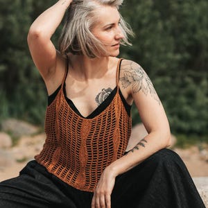 May include: A rust-colored sleeveless crochet tank top featuring a vertical ribbed pattern and thin spaghetti straps, worn over a black camisole paired with loose-fitting black trousers. The top has a V-neckline and an open-knit texture throughout.