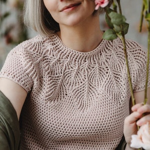 May include: A close-up of a pale pink crocheted top with a delicate leaf pattern around the neckline. The top has short sleeves and a relaxed fit.