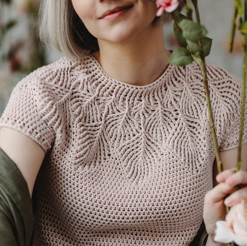 May include: A close-up of a pale pink crocheted top with a delicate leaf pattern around the neckline. The top has short sleeves and a relaxed fit.