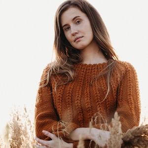 May include: A woman wearing a brown crocheted sweater stands in a field of tall grass. She is looking at the camera with a serious expression.