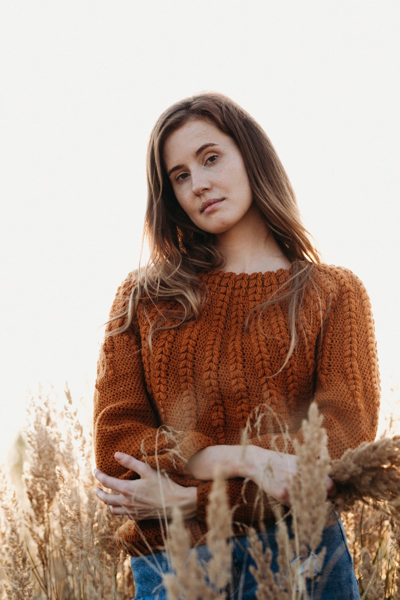 May include: A woman wearing a brown crocheted sweater stands in a field of tall grass. She is looking at the camera with a serious expression.