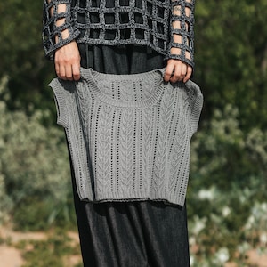 May include: A grey sleeveless knit top featuring a vertical chevron pattern and a round neckline. The garment has a ribbed collar and armholes, with a textured stitch design throughout the front panel, showcasing a consistent knit construction.