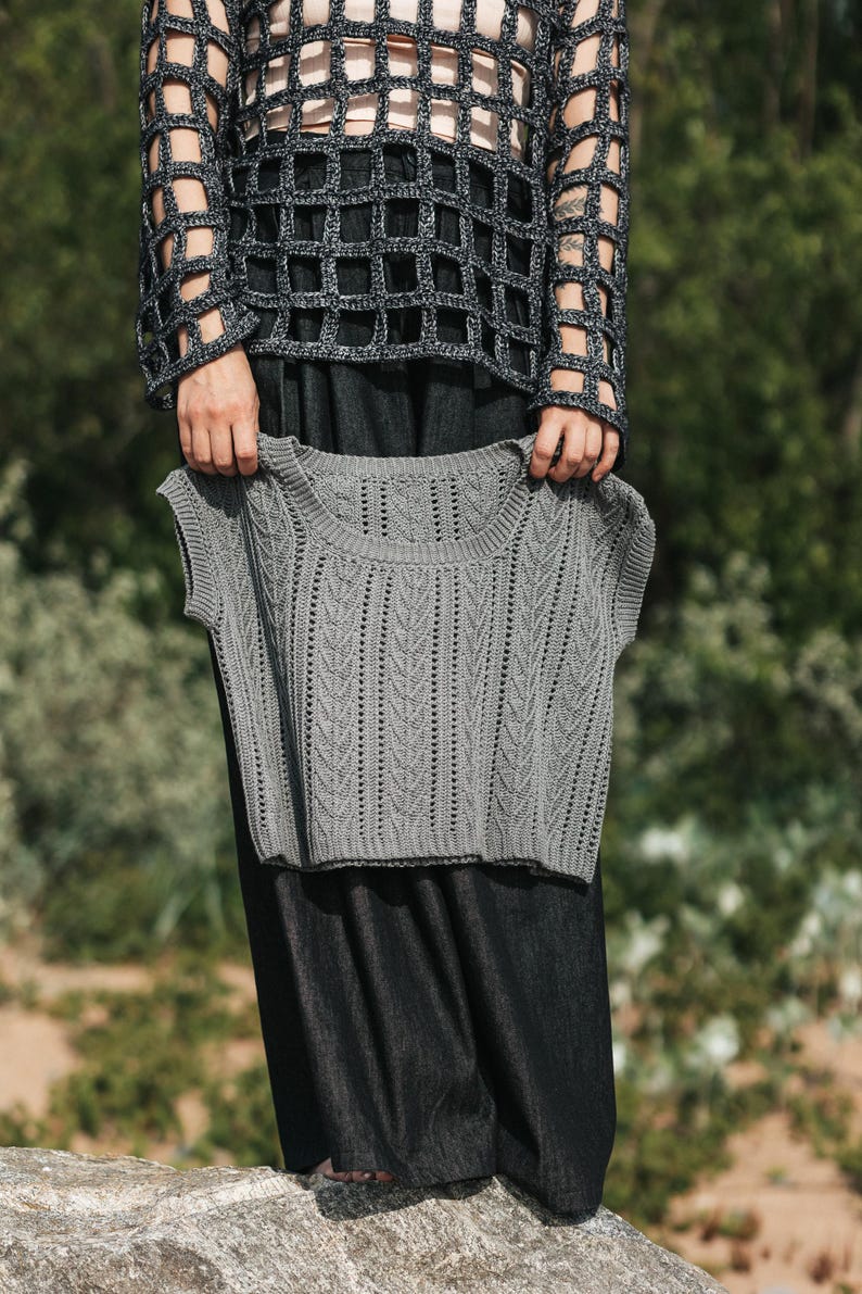 May include: A grey sleeveless knit top featuring a vertical chevron pattern and a round neckline. The garment has a ribbed collar and armholes, with a textured stitch design throughout the front panel, showcasing a consistent knit construction.