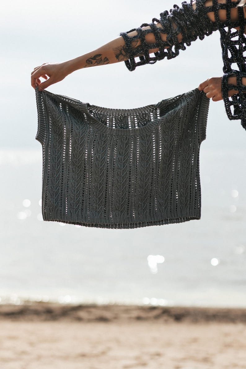 May include: A dark gray knit top featuring a vertical chevron stitch pattern, a wide round neckline, and short sleeves. The garment is constructed with a textured, open-work design that creates a subtle repeating pattern across the entire surface.