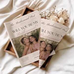May include: Two newspaper-style announcements with the text "WILL YOU BE MY Bridesmaid?" are displayed. The announcements feature a photo of three women and are accompanied by a bouquet of flowers in a wooden tray, all set against a white fabric backdrop.