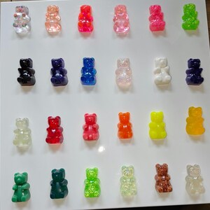 Puede incluir: Una colección de dijes de resina con forma de ositos de goma de colores. Los dijes son de varios colores, incluyendo rojo, rosa, verde, azul y amarillo. Algunos tienen purpurina o detalles florales. Los osos están dispuestos sobre una superficie blanca.