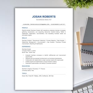 May include: A business analyst resume with the name "JOSAN ROBERTS" in blue text. The resume includes sections for profile, skills, experience, education, and tools. The resume is on a white background with a notebook and keyboard in the background.
