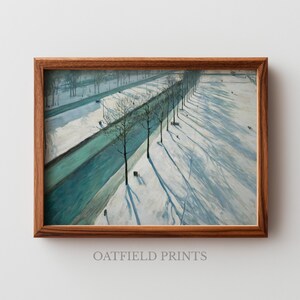 Op de afbeelding: Een ingelijst winterlandschapsschilderij met een bruine houten lijst. Het kunstwerk toont een besneeuwde scène met een turquoise waterweg, kale bomen en lange schaduwen. De tekst "OATFIELD PRINTS" staat onderaan.