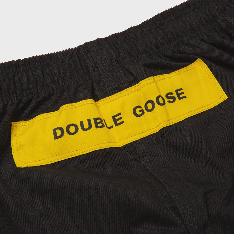 May include: Close-up of black athletic shorts with a yellow rectangular patch. The patch features the text "DOUBLE GOOSE" in black capital letters. The shorts have an elastic waistband and visible stitching.