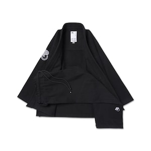 Op de afbeelding: Zwart vechtsportuniform, bestaande uit een jas en broek. De jas heeft een kraag en een wit logo op de linkermouw. De broek heeft een taille met trekkoord. Een wit logo staat ook op het rechteronderbeen.