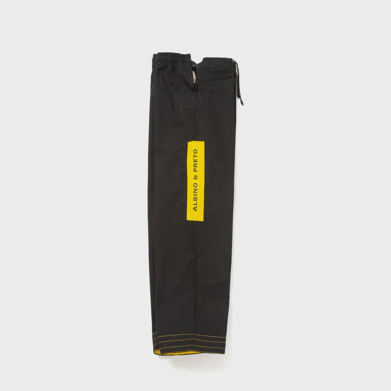 May include: Black martial arts pants with a yellow vertical stripe that reads "ALBINO & PRETO." The pants have yellow stitching along the bottom hem. The pants are folded and tied at the top with a drawstring.