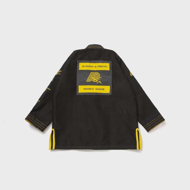May include: Black martial arts gi with yellow stitching and accents. The back features a rectangular patch with the text "ALBINO & PRETO" above a logo and the words "DOUBLE GOOSE" below. The gi has a traditional collar and sleeve design.
