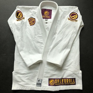 May include: White Jiu-Jitsu gi with the brand name "Shoyoroll" in gold and purple. The gi has patches on the shoulders and chest. The gi is laid out on a black surface.