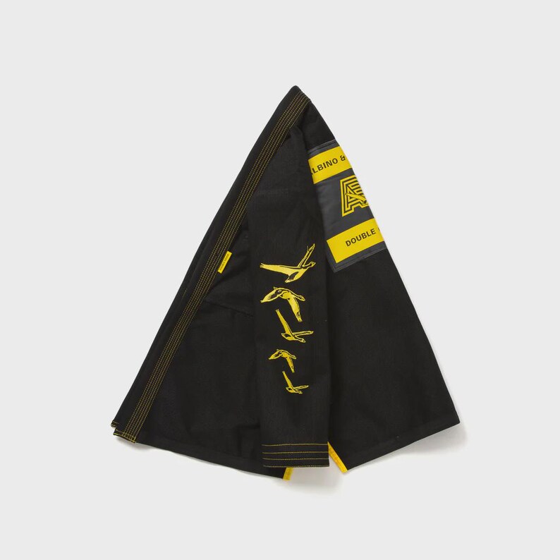 May include: Black martial arts uniform with yellow accents. The uniform features a yellow patch with the text "ALBINO & DOUBLE" and a stylized logo. The sleeve has a series of yellow bird graphics.