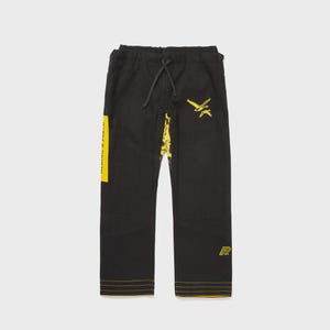 May include: Black martial arts pants with yellow accents. The pants feature a yellow stripe with text on the left leg, a bird graphic, and a logo. The bottom hem and seams are also yellow.