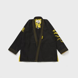 May include: Black martial arts gi with yellow accents. The gi features yellow stitching, sleeve details, and a yellow interior lining. A logo is visible on the chest and sleeve. The gi is laid flat on a light gray surface.