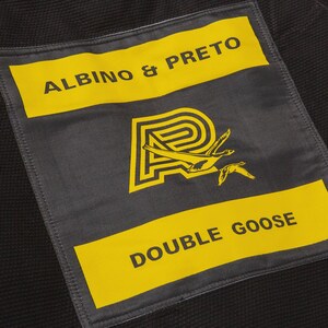 May include: Close-up of a black fabric patch with yellow text and a logo. The text reads "ALBINO & PRETO" and "DOUBLE GOOSE". The logo features a stylized "P" with two flying geese in yellow.