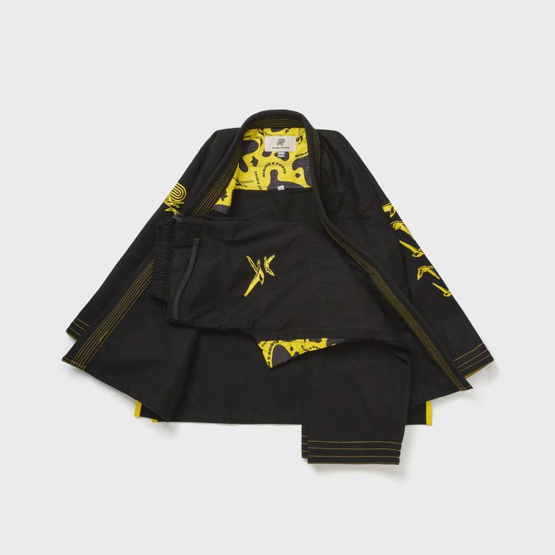 May include: Black martial arts uniform with yellow accents. The jacket and pants feature yellow stitching and decorative patterns. The interior lining of the jacket is yellow with a unique design. The uniform is laid out on a light gray surface.