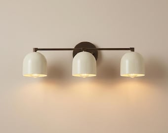May include: A three-light vanity fixture with a dark bronze finish. The fixture features three off-white, dome-shaped shades. The lights are mounted on a horizontal bar and a circular backplate. The lights are illuminated, casting a warm glow.