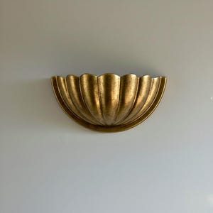 Könnte beinhalten: Eine goldfarbene Wandleuchte mit einem muschelförmigen Design. Die Leuchte hat eine strukturierte Oberfläche und ist an einer hellgrauen Wand befestigt. Das dekorative Stück ist aus Metall und hat eine halbkreisförmige Form.