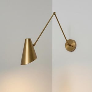 May include: A brass-toned wall-mounted lamp with an adjustable arm and conical shade. The lamp is mounted on a white wall, showcasing a minimalist design. The shade is angled downward, suggesting focused illumination.