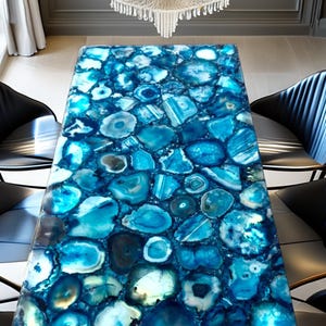 Op de afbeelding: Een rechthoekige tafel met een oppervlak van blauwe agaatstenen. De stenen variëren in grootte en vorm, waardoor een mozaïekachtig patroon ontstaat. De tafel is omringd door zwarte stoelen.