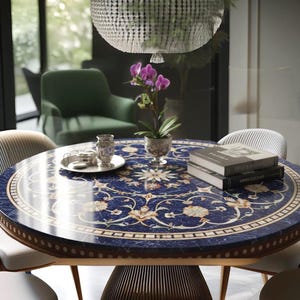 Puede incluir: Una mesa de comedor redonda con una superficie azul oscuro estampada, adornada con motivos florales y geométricos. La mesa tiene un borde y una base de madera clara, con una planta de orquídeas y libros. La mesa está rodeada de sillas.