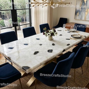 May include: A large, rectangular white quartz dining table with dark gray and black accents. The table is supported by gold-colored legs and surrounded by navy blue velvet chairs. A floral arrangement sits on the table.