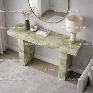 Puede incluir: Una consola de mármol verde con una parte superior rectangular y dos patas de soporte. La mesa está adornada con una lámpara, un jarrón con flores secas, un cuenco pequeño y libros. Un espejo redondo cuelga de la pared. La mesa mide unos 150 cm de largo.