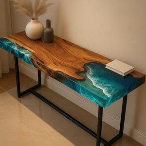 Puede incluir: Una mesa de madera larga y rectangular con un borde vivo y un diseño de río de resina turquesa. La mesa tiene patas de metal negro y está rematada con un pequeño libro blanco. Un jarrón y una botella están sobre la mesa.
