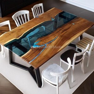 Könnte beinhalten: Rechteckiger Esstisch aus Holz mit blauem Harzfluss-Design. Der Tisch hat ein dunkles Metallgestell und ist von weißen Stühlen umgeben. Die Tischmaße betragen ca. 183 cm Länge und 91 cm Breite.