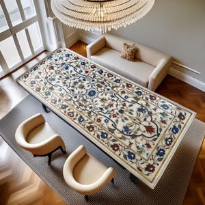 Puede incluir: Una mesa rectangular de mármol con intrincados incrustaciones florales en tonos rojos, azules y verdes. La mesa está rodeada por dos sillas color crema y un sofá color crema. Una gran lámpara de araña cuelga sobre la mesa, iluminando la habitación.