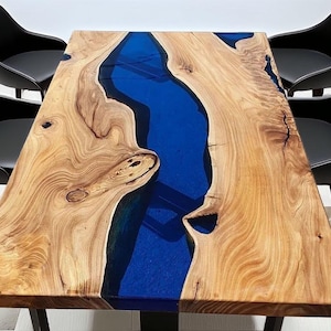 Op de afbeelding: Houten eettafel met een blauwe epoxy rivier in het midden. Vier zwarte stoelen met een modern design omringen de tafel. De houtnerf is zichtbaar en de tafel staat op zwarte metalen poten. Afmetingen: ongeveer 150 cm x 80 cm.