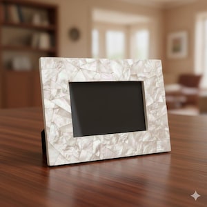 Puede incluir: Marco de fotos rectangular con diseño de mosaico. El marco es blanco con detalles iridiscentes, y el interior es negro. Se muestra sobre una superficie de madera marrón oscuro, un artículo de decoración.