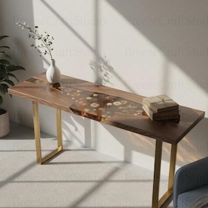 Op de afbeelding: Een houten console met een heldere harsinleg gevuld met gedroogde bloemen. De tafel heeft een donkere houten afwerking, gouden metalen poten en een kleine stapel boeken. Een witte vaas met een plant staat ook op de tafel.