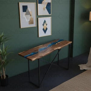 Puede incluir: Una consola de madera con un diseño de río de resina azul. La mesa tiene un borde de madera natural y se apoya en patas de metal negro. La habitación tiene una pared verde oscuro, obras de arte, un sillón azul y una lámpara de pie.