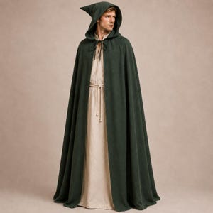 Puede incluir: Una capa con capucha de color verde bosque oscuro que llega hasta el suelo, hecha de tela sólida, con capucha puntiaguda y cierre de lazo frontal. Debajo de la capa hay una túnica larga de color beige claro con cuello redondo y un cordón.