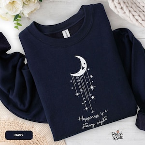 May include: Navy blue sweatshirt featuring a white crescent moon and stars design. The text "Happiness is a starry night" is printed below the design. The sweatshirt is from Peach & Salt.