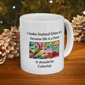 May include: White ceramic mug with the text "I make Stained Glass art because life is a Pane it should be Colorful." The mug features a colorful stained glass design. The mug is on a wooden surface.