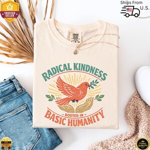 May include: Cream-colored t-shirt with the words "RADICAL KINDNESS" and "ROOTED IN BASIC HUMANITY" in green and orange. A red bird with a green olive branch is in the center. The shirt is folded on a white surface with blue jeans.