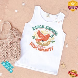 May include: White tank top with the text "RADICAL KINDNESS ROOTED IN BASIC HUMANITY" and a graphic of a red bird holding an olive branch. A beige woven tote bag is on the left. The background is a sandy surface.