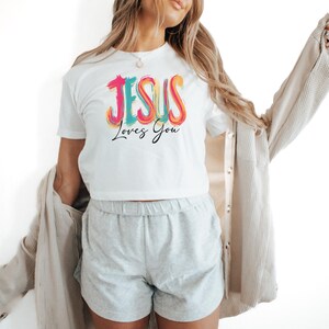 May include: White t-shirt with the text "JESUS Loves You" in colorful, stylized lettering. The shirt is short-sleeved and appears to be made of a soft material. The text is in pink, teal, orange, and yellow.