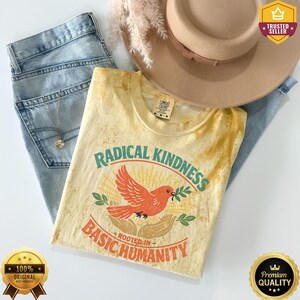 May include: A beige t-shirt with a distressed texture featuring a graphic of a red bird flying above two hands, surrounded by the text Radical Kindness Rooted In Basic Humanity. The shirt is paired with light blue denim jeans and a tan hat.