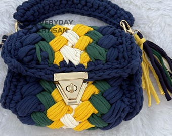 May include: A navy blue, hand-crocheted handbag with a woven pattern of yellow, green, and white. The bag features a gold-colored clasp, a braided handle, and a tassel. The text "EVERYDAY ARTISAN" is visible on the top of the bag.