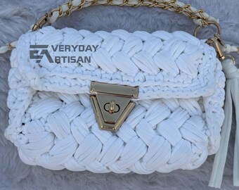 May include: A white, hand-crocheted handbag with a gold-tone clasp and chain strap. The bag features a braided design and a matching tassel. The text "VERYDAY ARTISAN" is visible on the front of the bag.