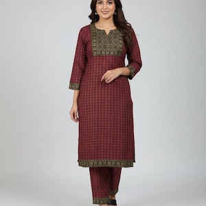 May include: A burgundy plaid kurta set. The top has a v-neck with a gold and black embroidered design. The sleeves and hem are trimmed with a matching gold and black border. The matching trousers also have a gold and black border.